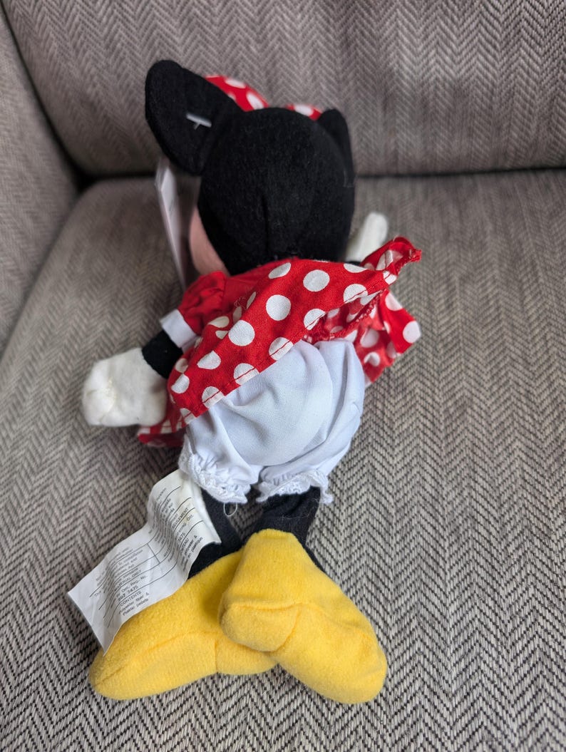K&ouml;nnte beinhalten: Ein Minnie Mouse Pl&uuml;schtier, mit einem roten Kleid mit wei&szlig;en Punkten, einer passenden Schleife und gelben Schuhen. Die schwarzen Ohren und das Gesicht sind sichtbar. Das Spielzeug liegt auf einer grauen Oberfl&auml;che.