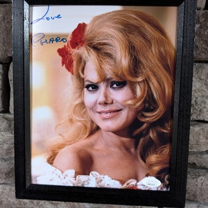 May include: A framed black and white photo of a woman with blonde hair and a red flower in her hair. The woman is smiling and looking at the camera. The photo is signed "Love,  [illegible]" in blue ink.