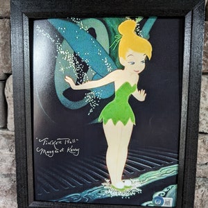 May include: A framed print of Tinker Bell, a fairy with green wings and a green dress, standing on a blue and white surface. The print is signed "Tinker Bell" and "Margaret Kerry".
