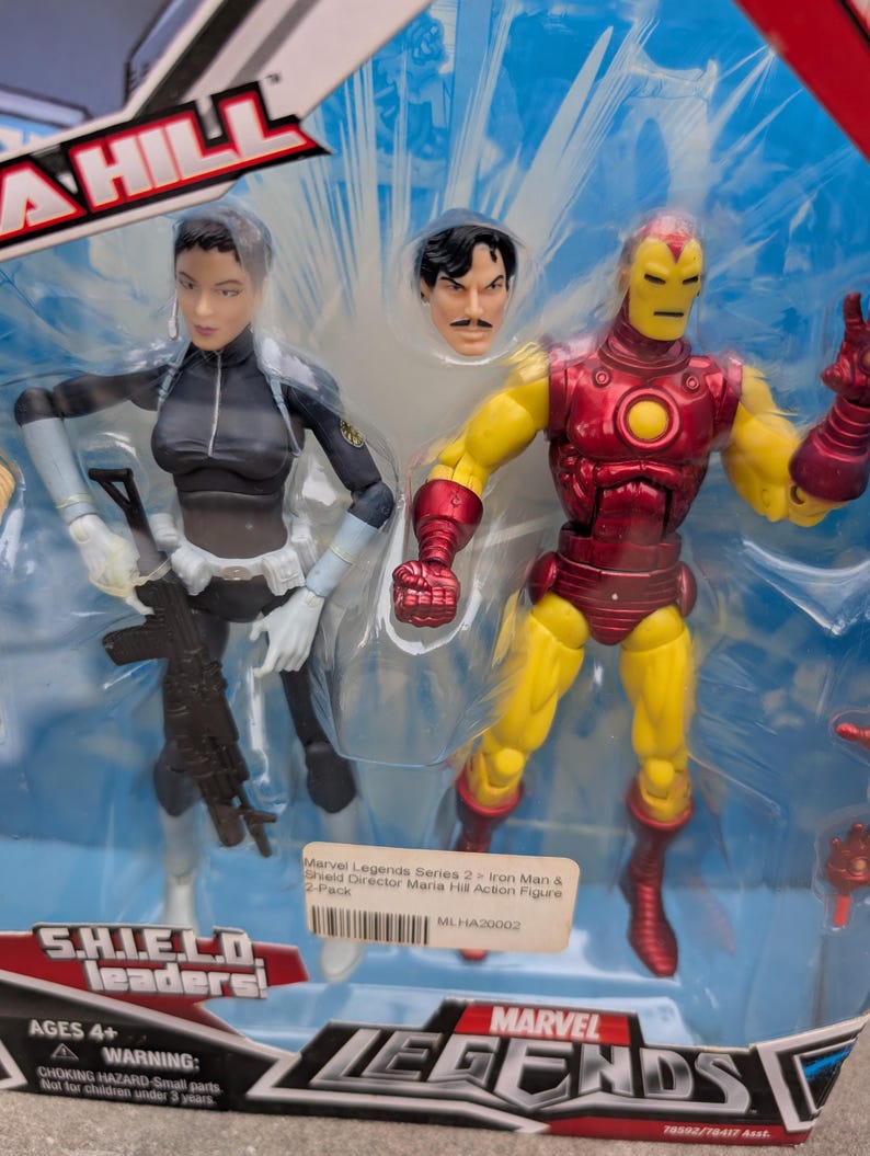 May include: A plastic package containing two action figures, one of Iron Man in a red and yellow suit and the other of Maria Hill in a black and white suit. The package is labeled "Marvel Legends Series 2" and "Iron Man & Shield Director Maria Hill Action Figure 2-Pack".
