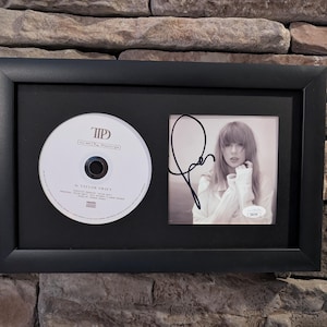 May include: A black framed display featuring a CD and a signed photograph of Taylor Swift. The CD is titled "1989 (Taylor's Version)" and the photograph is from the "Red (Taylor's Version)" album.
