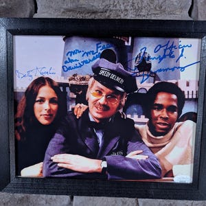 May include: A framed photograph of three people, two women and one man, with their autographs. The man is wearing a blue uniform with the words "Speedy Delivery" on his hat. The women are wearing casual clothing. The background is a cartoon-like cityscape.