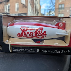 May include: A die-cast metal replica of a Pepsi-Cola blimp, featuring a red and white color scheme with the Pepsi logo. The blimp is presented in a box with the text "Blimp Replica Bank" and "Limited Edition".