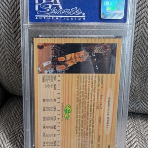 May include: A sports trading card encased in a clear plastic holder. The card features a photo of Shaquille O'Neal in a basketball uniform. The back of the card includes statistics and biographical information. The holder is labeled "Professional Sports Authenticator."