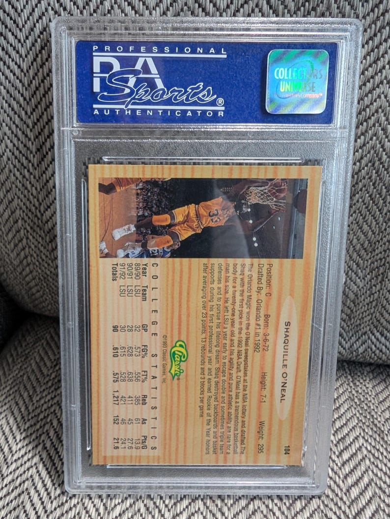 May include: A sports trading card encased in a clear plastic holder. The card features a photo of Shaquille O'Neal in a basketball uniform. The back of the card includes statistics and biographical information. The holder is labeled "Professional Sports Authenticator."