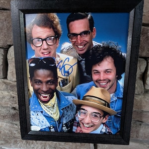 May include: A framed photograph featuring a group of five smiling people. The photo is in a black frame and appears to be an autographed print. The subjects are wearing glasses and denim jackets, and one is wearing a hat.