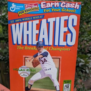 Op de afbeelding: Een oranje doos Wheaties-ontbijtgranen met een honkbalthema. De doos heeft de tekst "WHEATIES" in grote witte letters. Het toont een afbeelding van honkbalspeler Nolan Ryan en de tekst "The Breakfast of Champions". De doos bevat 340g ontbijtgranen.