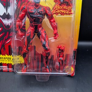 May include: A red and black action figure of the character Carnage from the Marvel Comics universe, packaged in a clear plastic blister pack. The figure is in a dynamic pose, with the words "MAXIMUM CARNAGE" visible on the packaging.