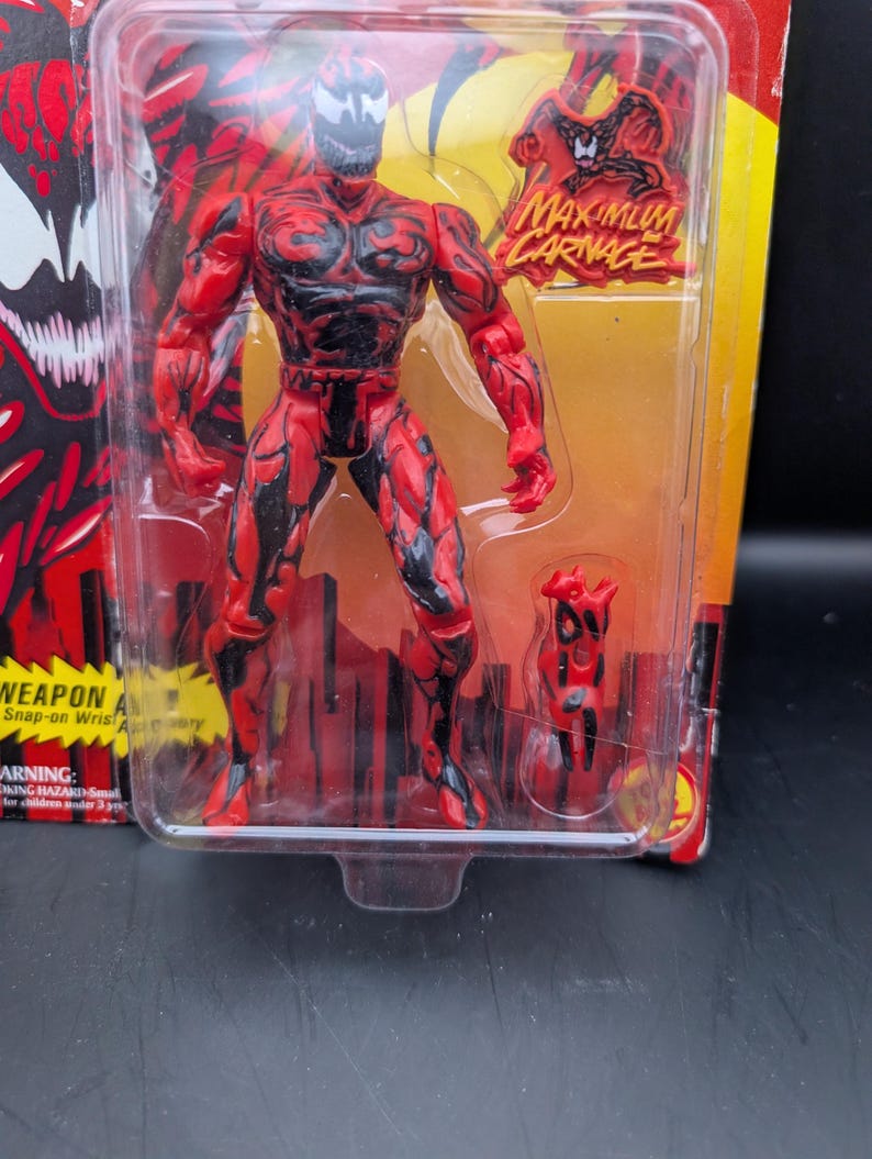 May include: A red and black action figure of the character Carnage from the Marvel Comics universe, packaged in a clear plastic blister pack. The figure is in a dynamic pose, with the words "MAXIMUM CARNAGE" visible on the packaging.