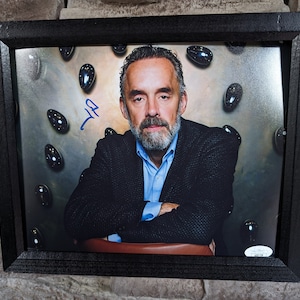 May include: A framed black and white photograph of a man with a beard and mustache. He is wearing a dark suit jacket and a light blue shirt. The background is a light brown color with black and silver objects scattered around. The man's signature is in blue ink in the top left corner of the photo.