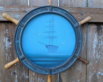 May include: A blue and black ship's wheel wall decor with a painted image of a ship on a blue background. The wheel has wooden spokes and the words "COOS BAY" and "BIRCH" are printed on the rim.
