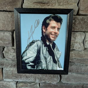 Puede incluir: Fotografía enmarcada de un hombre sonriente con cabello oscuro, vistiendo una chaqueta de cuero negra. La foto está sobre un fondo azul claro y está firmada. El marco es negro y rectangular.