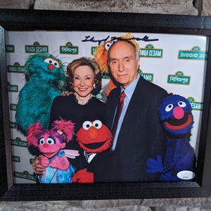 May include: A framed photo of a man and woman posing with Sesame Street characters. The characters include Big Bird, Elmo, and Cookie Monster. The man and woman are smiling and holding the puppets. The photo is signed by the man.