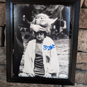 May include: A black and white photograph of a young person wearing a white robe with a green alien character on top of their head. The photo is framed in a black frame and has a blue signature in the bottom right corner.