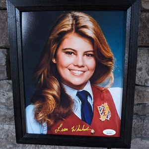Puede incluir: Una fotografía enmarcada de una mujer con cabello largo y castaño que lleva un chaleco rojo con una corbata azul y un broche dorado. La mujer sonríe y mira a la cámara. La foto está firmada "Lisa Whelchel" con tinta dorada.