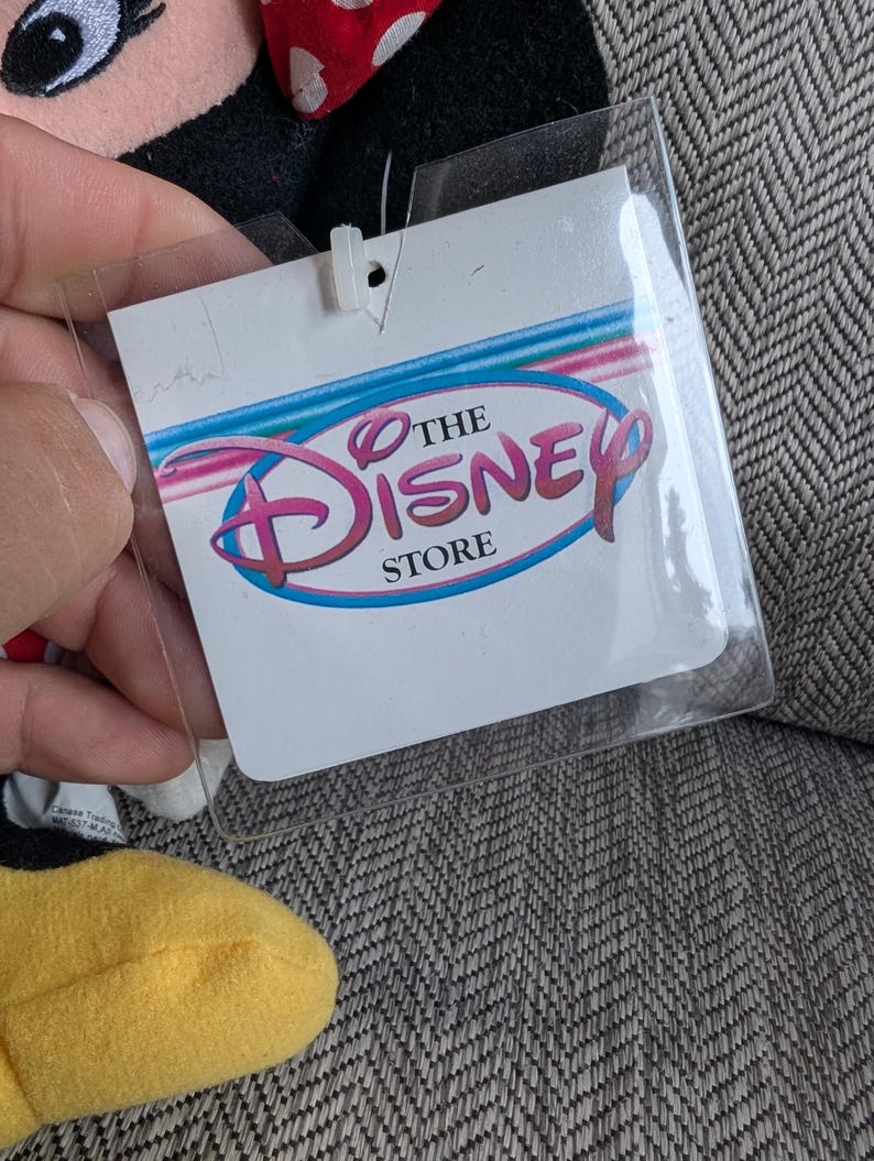 K&ouml;nnte beinhalten: Ein durchsichtiges Kunststoffetikett mit den Worten "THE DISNEY STORE" in Rosa und Blau, befestigt an einem Pl&uuml;schtier. Das Etikett ist rechteckig mit abgerundeten Ecken und einem kleinen Loch oben. Das Disney-Logo ist deutlich sichtbar.