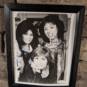 May include: Framed black and white photograph of three women, with blue ink signatures. The photograph is in a black frame and mounted on a white background. The women are smiling.