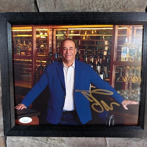 May include: Framed photograph featuring a man in a blue blazer and white shirt, standing in front of a bar with bottles and glasses. The photograph is signed in gold ink. The frame is black.
