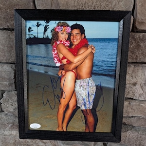 May include: Framed photograph of a couple embracing on a beach. The woman wears a red bikini and floral headpiece, the man wears patterned swim trunks and a red lei. The photo is signed and framed in a black wooden frame.