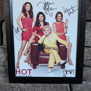 May include: A framed photograph of the cast of the TV show "Hot in Cleveland". The photograph features Betty White, Valerie Bertinelli, Jane Leeves, and Wendie Malick. Each actress has signed their name in black ink. The photograph is set against a white background and features the show's logo and title.