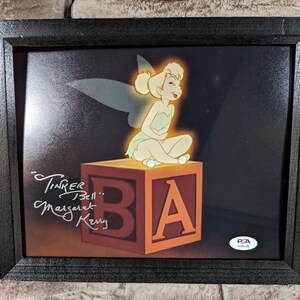 May include: Framed artwork featuring Tinker Bell sitting on a block with the letter "A". The artwork is signed "Tinker Bell" and "Margaret Kerry". The frame is black.
