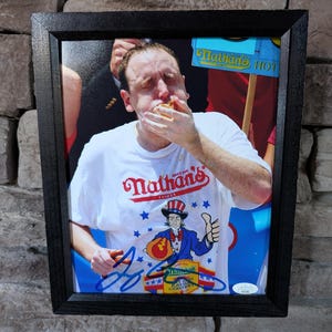 以下が含まれることがあります： ホットドッグを食べている男性の写真が額装されており、Nathan's Famousのロゴと漫画のアンクルサムのデザインが入った白いTシャツを着ています。写真はサイン入りで、レンガの壁に飾られています。