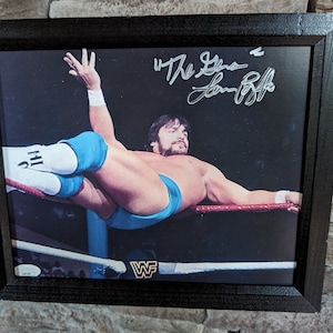 Puede incluir: Una foto en blanco y negro de un luchador con un traje de lucha azul y blanco, suspendido boca abajo en un ring de lucha. El luchador lleva calcetines blancos con las letras "2HH" en ellos. La foto está firmada "The Great" y "Lex Luger".