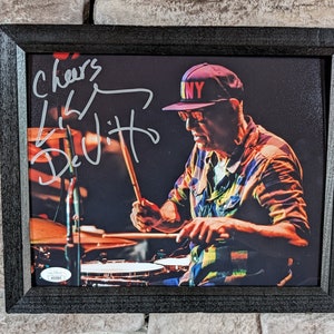 Può includere: Una foto in bianco e nero incorniciata di un uomo che suona la batteria. Indossa una camicia a quadri e un cappello nero con la scritta "NY" sopra. La foto è firmata "Cheers,  Little DeVitts".