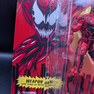May include: A red and black action figure of a character with a white face and sharp teeth, packaged in a clear plastic case. The packaging is red with a cityscape design and text that reads "WEAPON ARM" and "WARNING: CHOKING HAZARD-Small Not for children under 3 yrs."