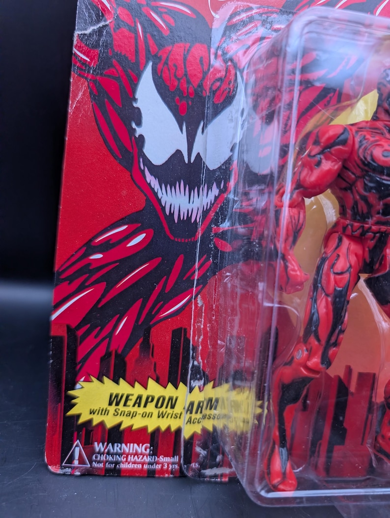 May include: A red and black action figure of a character with a white face and sharp teeth, packaged in a clear plastic case. The packaging is red with a cityscape design and text that reads "WEAPON ARM" and "WARNING: CHOKING HAZARD-Small Not for children under 3 yrs."
