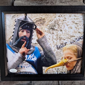 以下が含まれることがあります： 映画「モンティ・パイソン・アンド・ホーリー・グレイル」の2人の俳優の写真が額装されています。俳優たちは中世の衣装を着ています。左側の俳優は青と白のチュニックとヘルメットを着ています。右側の俳優は茶色のチュニックを着ており、顔に偽の鼻をつけています。写真は両方の俳優によって「Best Wishes」というメッセージと共に署名されています。