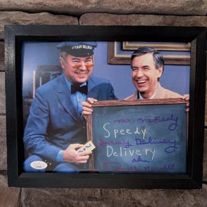 May include: Framed photograph featuring two smiling men, one in a blue uniform and cap, the other in a tan jacket. A chalkboard reads "Speedy Delivery" with signatures. The frame is black, and the background is a brick wall.