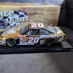Op de afbeelding: Een goud-zwarte gegoten modelraceauto met het nummer 20 op de zijkant en het dak. De auto heeft sponsorlogo's en wordt op een zwarte basis gepresenteerd. Een doos met "Race Fans" staat op de achtergrond.