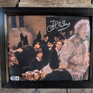 以下が含まれることがあります： ハリーポッター映画シリーズのシーンが描かれた額装されたプリント。食堂にいる生徒たちのグループが描かれている。プリントには銀色のインクで「J.K. ローリング」のサインが入っている。