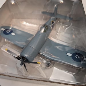 May include: A grey die-cast model aeroplane in a clear plastic case. The wings are blue with white and blue circular markings. The propeller has black blades with yellow tips. This model aeroplane is a collector's item.