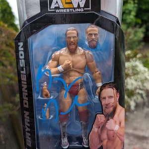 Könnte beinhalten: Eine Bryan Danielson Actionfigur aus der AEW Unmatched Collection, Serie 5, in einer schwarzen und transparenten Kunststoffverpackung. Die Figur ist ein muskulöser Mann mit Bart, der rote und weiße Wrestling-Kleidung trägt. Enthält einen zusätzlichen Kopf und Hände.