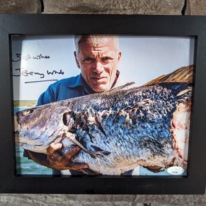 May include: Framed photograph featuring a man holding a large fish. The fish has blue and silver scales. The photograph is signed "Jeremy Wade" with the inscription "Best wishes". The frame is black and the background is a brick wall.
