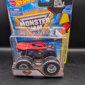 May include: A Hot Wheels Monster Jam truck featuring a Superman design. The blue truck has large black wheels and a red cape. The packaging includes the Hot Wheels and Monster Jam logos, and the text "Includes Snap-On Battle Slammer!"