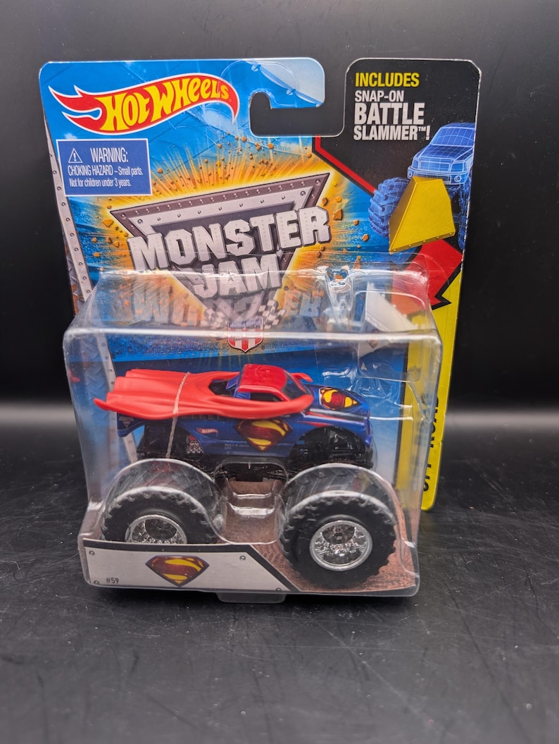 May include: A Hot Wheels Monster Jam truck featuring a Superman design. The blue truck has large black wheels and a red cape. The packaging includes the Hot Wheels and Monster Jam logos, and the text "Includes Snap-On Battle Slammer!"