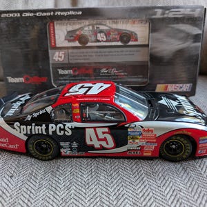 Op de afbeelding: Een 1:24 schaal gegoten replica van een NASCAR raceauto. De auto is voornamelijk zwart, rood en zilver, met het nummer 45 prominent aanwezig. Het heeft sponsorlogo's en de tekst "Sprint PCS". De doos vermeldt "2001 Die-Cast Replica".