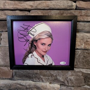 May include: Framed photograph of a woman in a nurse's uniform, with a white cap and a white uniform with black trim. The photo has an autograph and is set against a purple background. The frame is black.