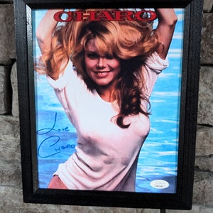 May include: A framed photo of a woman with long blonde hair wearing a white shirt. The woman is smiling and looking at the camera. The photo is signed "Love Charo" in blue ink. The photo is framed in a black frame.