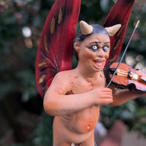 May include: A figurine of a devil-like creature with red wings, horns, and a violin. The figure is nude with a light skin tone and is playing the violin. The wings are red with black and gold accents. The background is blurred green.