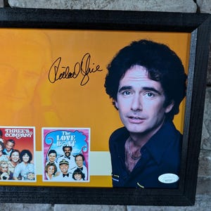May include: A framed photograph of actor Robert Guillaume, signed with his name. The photo shows him wearing a brown jacket and a white shirt. The photo also includes two smaller photographs of the TV shows "Three's Company" and "The Love Boat".