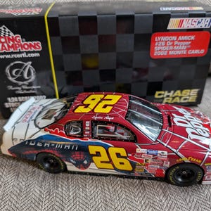 Op de afbeelding: Een 1:24 schaalmodel van een Chevrolet Monte Carlo uit 2002, in rood, wit en zwart geverfd. De auto is voorzien van Dr Pepper-branding en heeft een Spider-Man-ontwerp met een rood spinnenwebpatroon en de tekst "Spider-Man" aan de zijkant. De auto staat op een witte achtergrond.