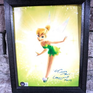 May include: Framed print of Tinker Bell, a Disney fairy with green wings and a green dress, against a yellow background with sparkles. The print is signed "Tinker Bell" and "Margaret Kerry".