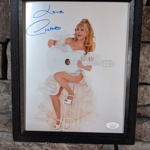 May include: A framed photograph of a woman in a white dress holding a white guitar. The woman is smiling and looking at the camera. The photo is signed "Love Hard" in blue ink.