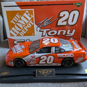 Op de afbeelding: Oranje en witte modelraceauto met nummer 20, gesponsord door The Home Depot en Ridgid. Het model staat op een zwart display met een gouden embleem. De doos erachter is oranje met nummer 20.