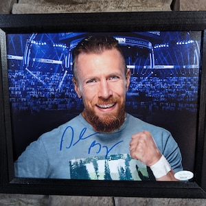 Könnte beinhalten: Ein gerahmtes Foto eines Mannes mit Bart, der lächelt und die Faust hebt. Er trägt ein blaues Hemd mit einem grün-weißen Muster. Das Foto ist mit einer blauen Unterschrift signiert. Der Hintergrund ist ein verschwommenes Bild eines Stadions mit einer Menschenmenge.