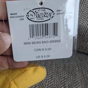 K&ouml;nnte beinhalten: Ein wei&szlig;es Etikett von The Disney Store f&uuml;r einen Minnie Mouse Mini-Beanbag. Das Etikett hat schwarzen Text und einen Barcode. Auf dem Etikett steht "MINI BEAN BAG MINNIE". Das Etikett befindet sich in einer transparenten Kunststoffh&uuml;lle.